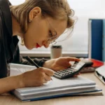 Mulher em uma mesa com documentos e uma calculadora, enquanto olha minuciosamente o documento.