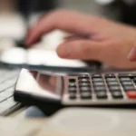 Mão tocando em um celular atrás de uma calculadora, exemplificando o cálculo do INSS Pró-labore